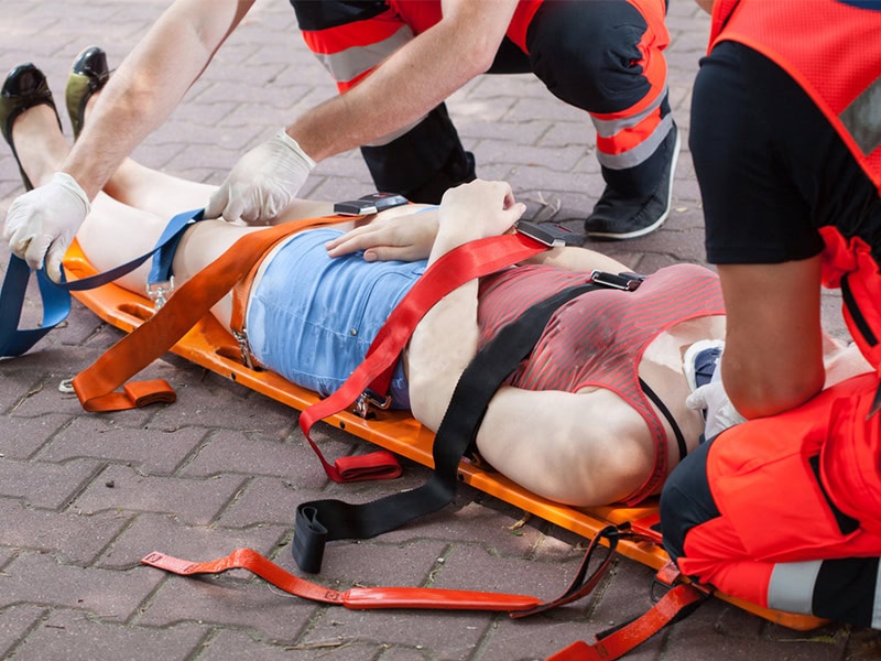 Emergency responders secure an injured woman to an orange spine board using multiple safety straps.