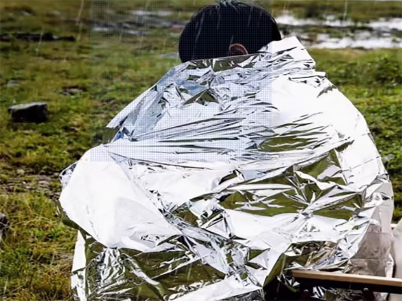 Person wrapped in a reflective emergency blanket outdoors, staying warm and dry in a cold, wet environment.