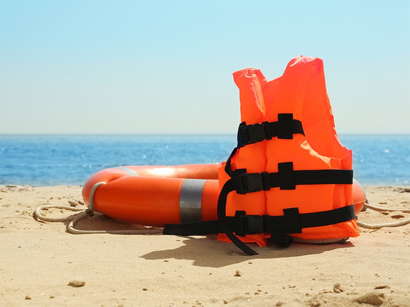 Bright orange life jacket and lifebuoy placed on sandy beach, ready for water safety and rescue.