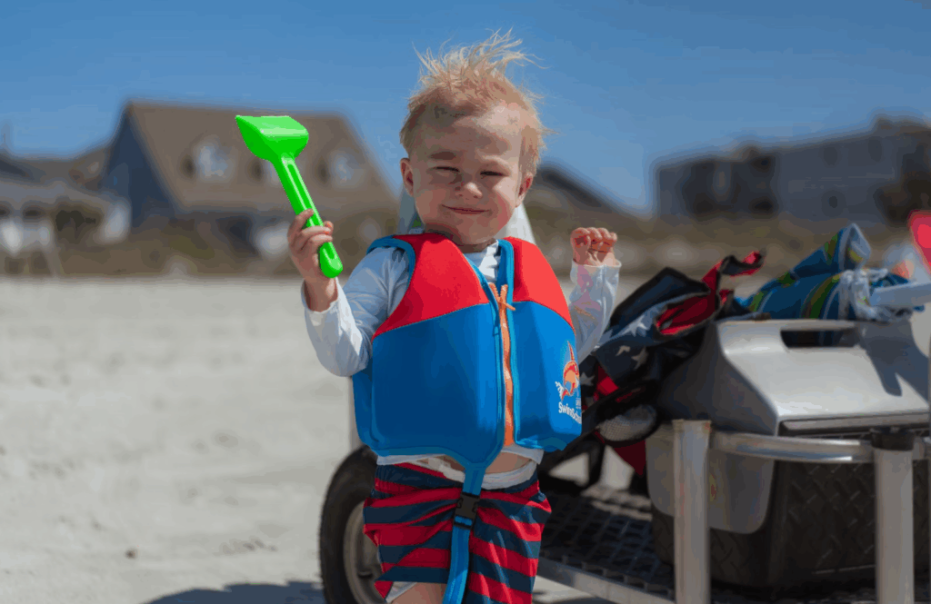 Smiling toddler in a colorful life jacket holds a toy shovel while enjoying a sunny day at the beach.