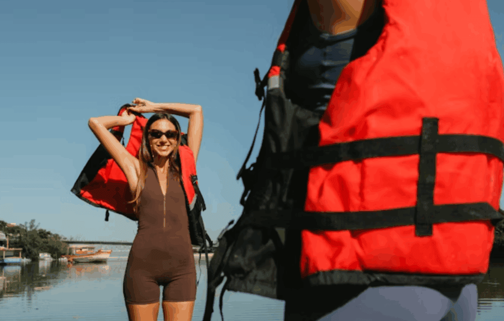 Smiling woman by the water prepares to put on a red life jacket, ready for a day of safe boating or kayaking.