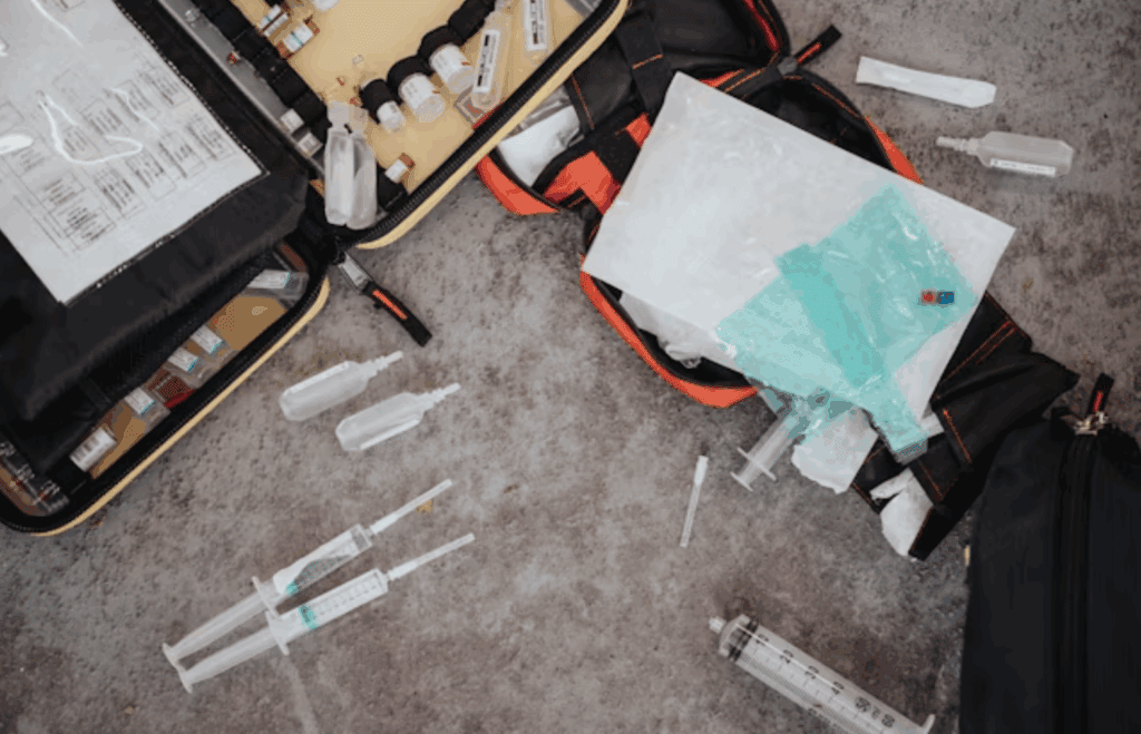 Open emergency medical kit with syringes, vials, and supplies scattered on the ground during urgent care.