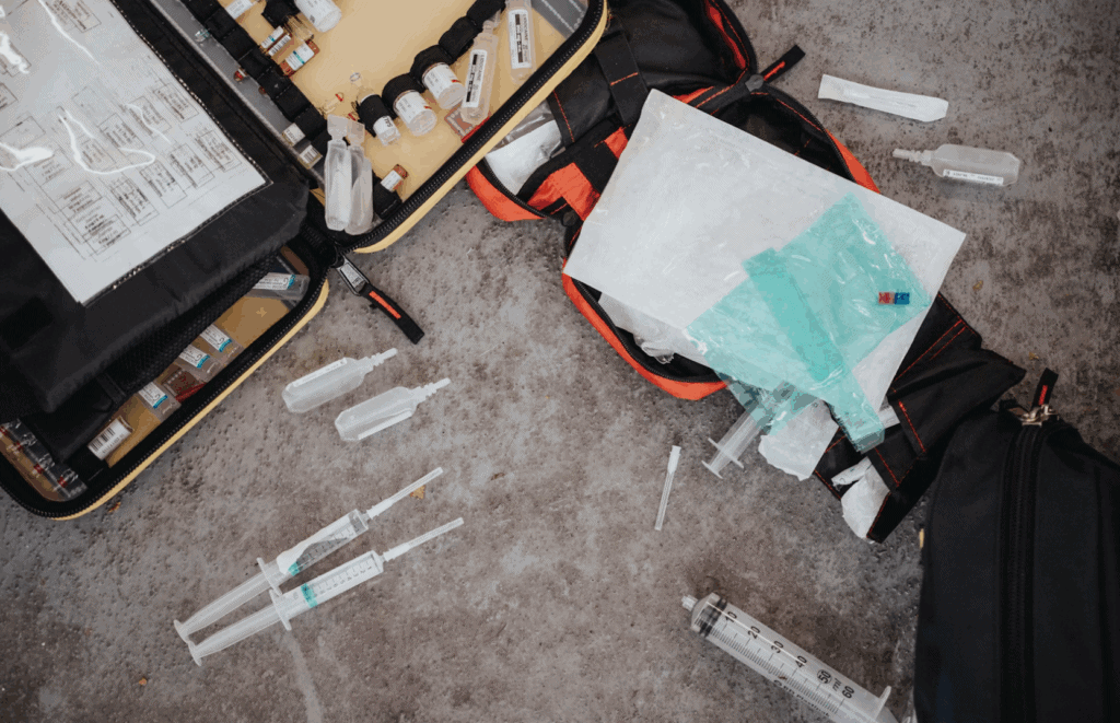 Open emergency medical kit with syringes, vials, and supplies scattered on the ground during urgent care.